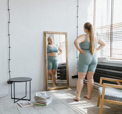 Plus size woman in sportswear stands barefoot, looking at her reflection in a wall mirror.